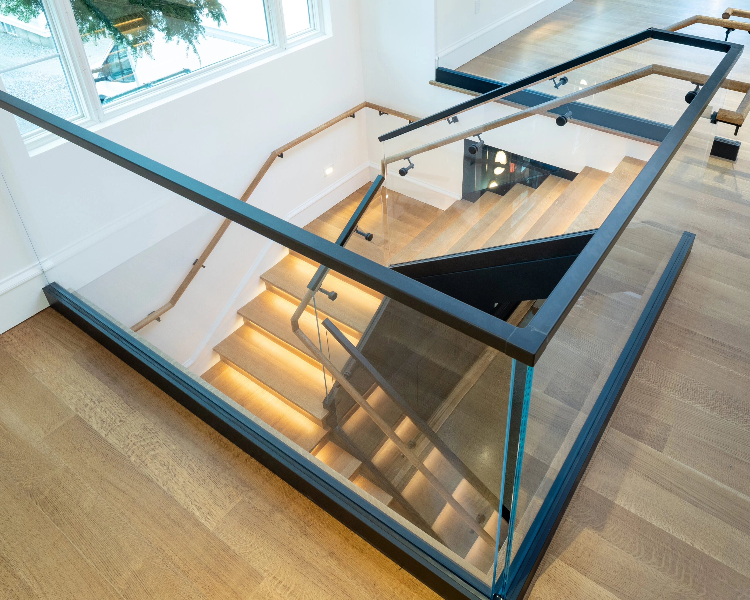 Looking down a stair where a wood-capped glass railing follows lit treads