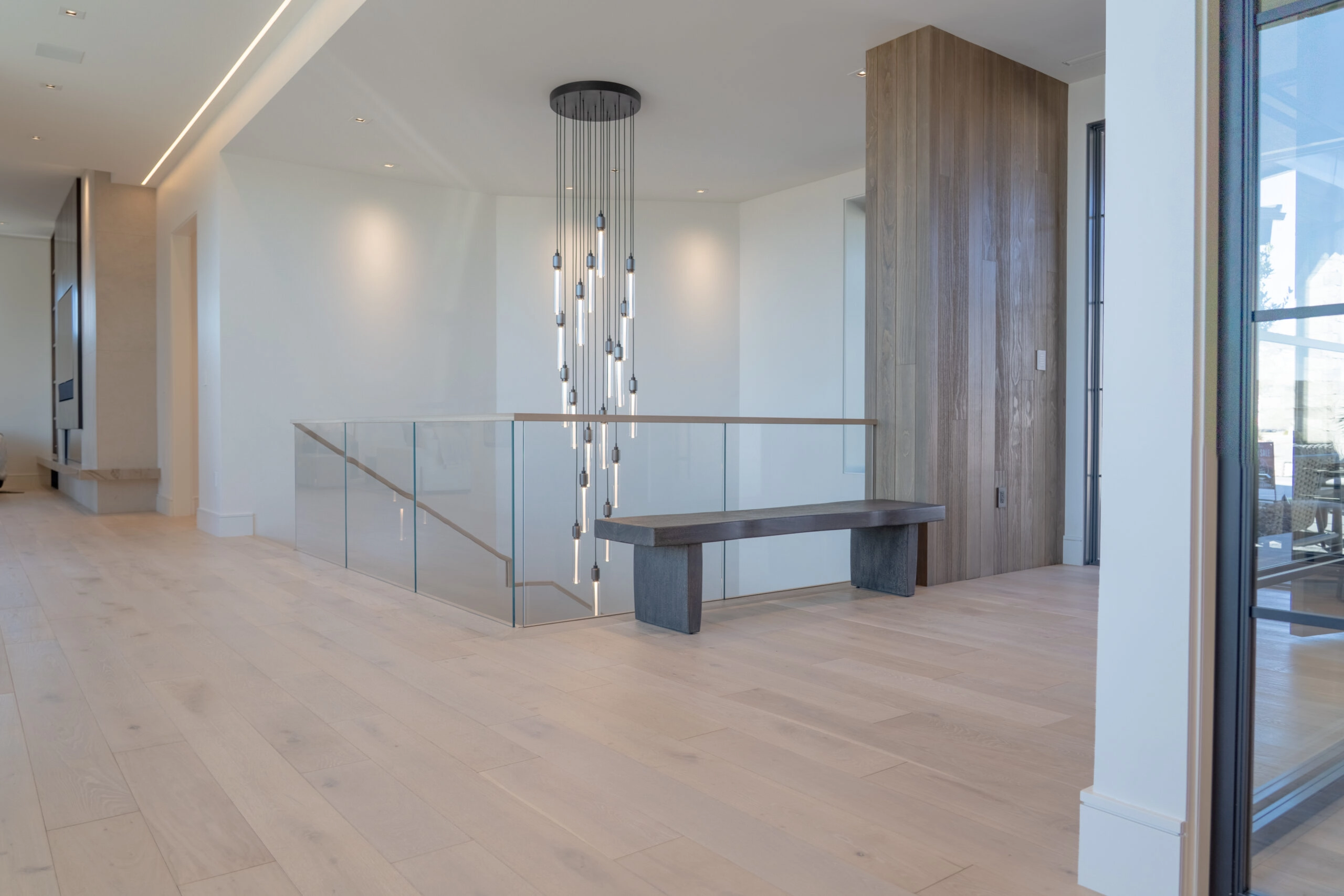 Glass railing edges a stairwell opening in a pale room lit by a linear chandelier