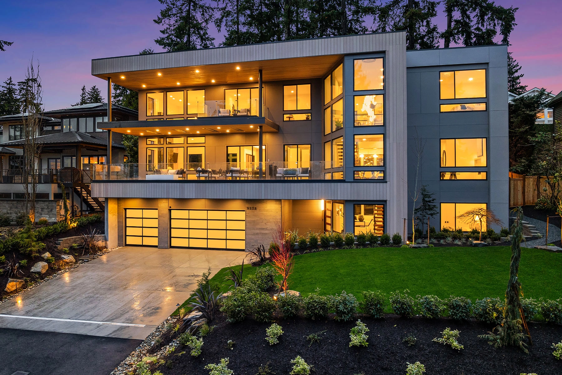 Illuminated hillside home at dusk with stacked balconies and glass railing above the driveway