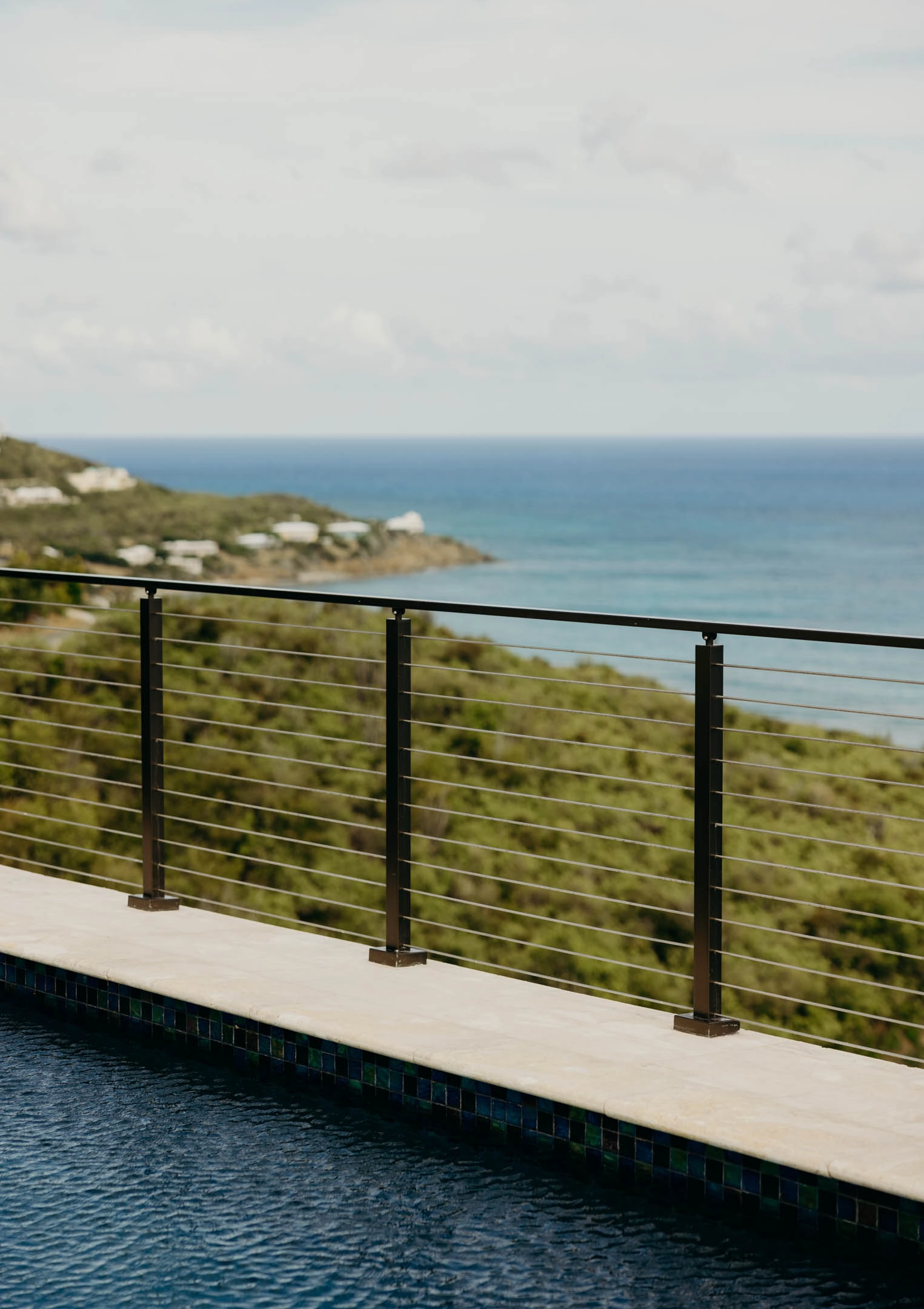 Black rod railing along a pool edge above a green hillside and ocean