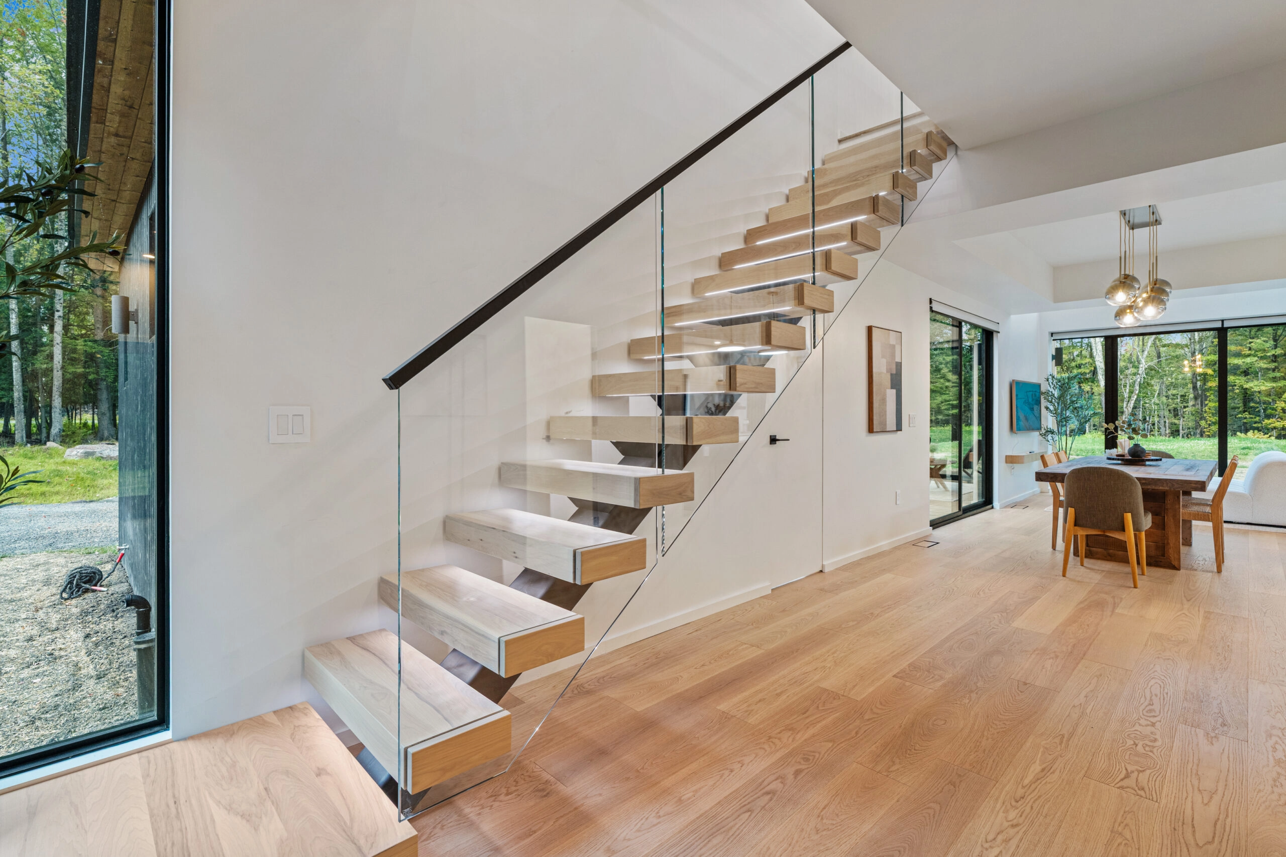 Floating stair with wood treads and glass railing rising beside a dining area