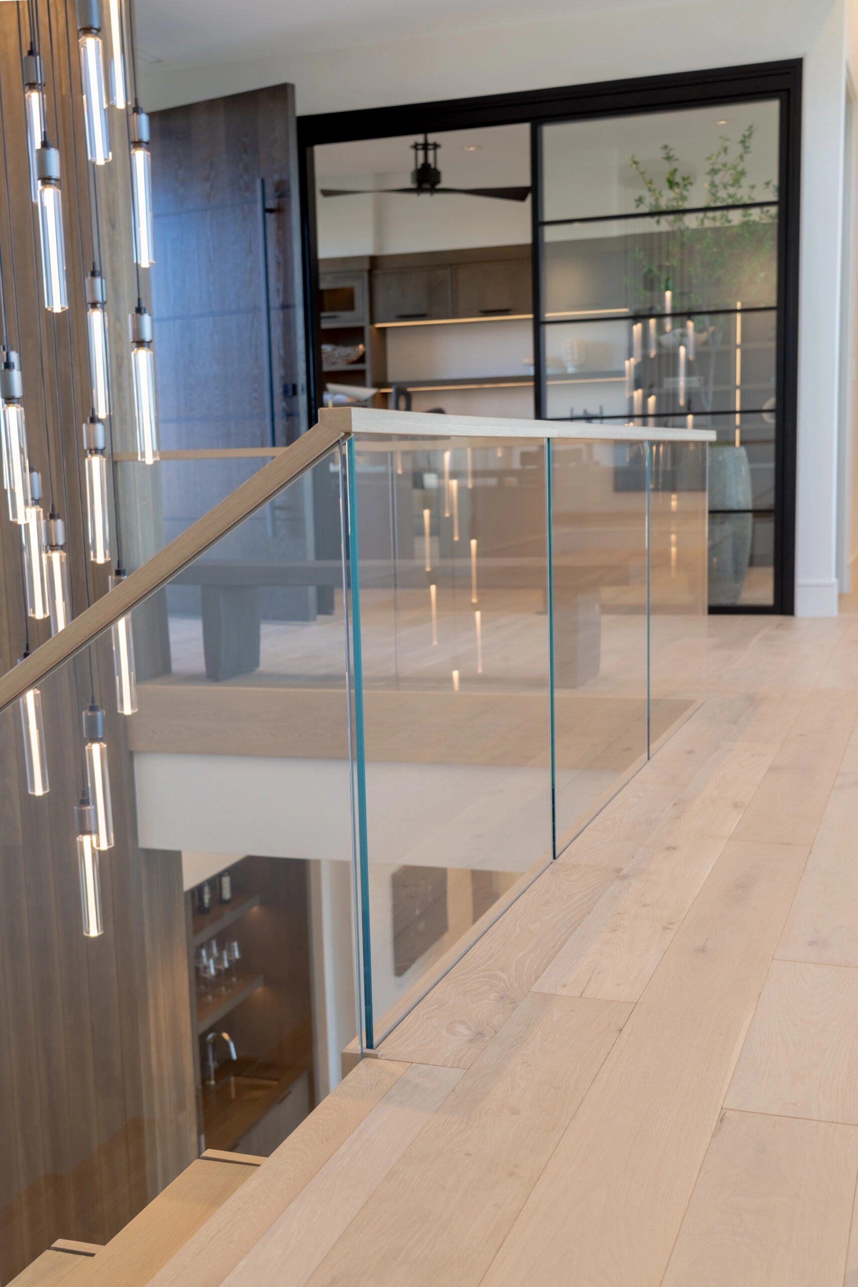 Glass railing lining a hallway beside dark paneling and a lit kitchen beyond