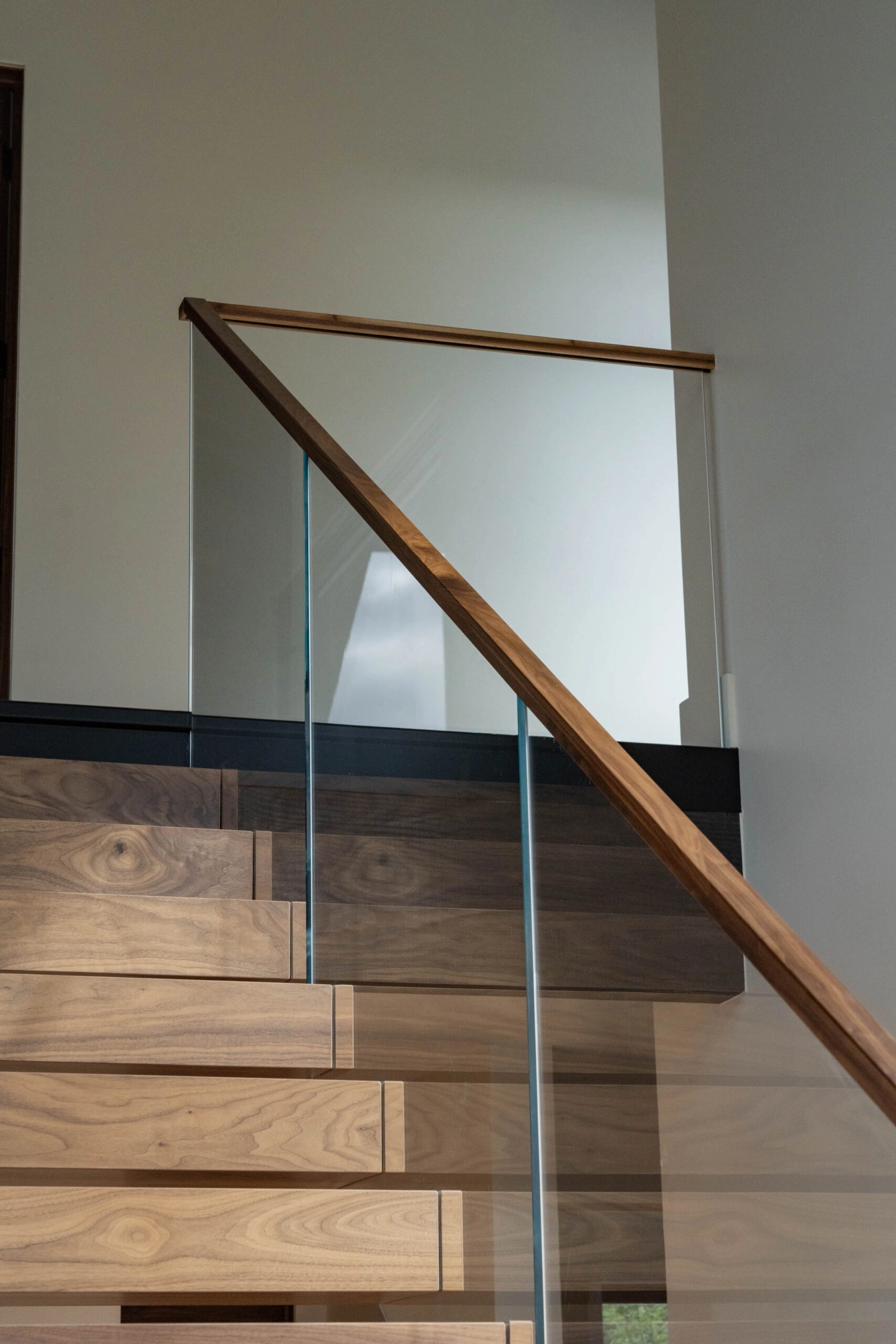 Wood handrail angling over a glass panel beside stair treads and a dark wall