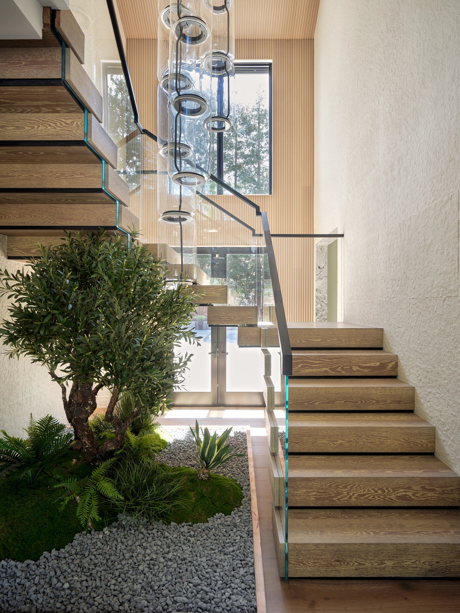 Wood box treads climb past glass railing above an indoor planting bed and cylinder pendants
