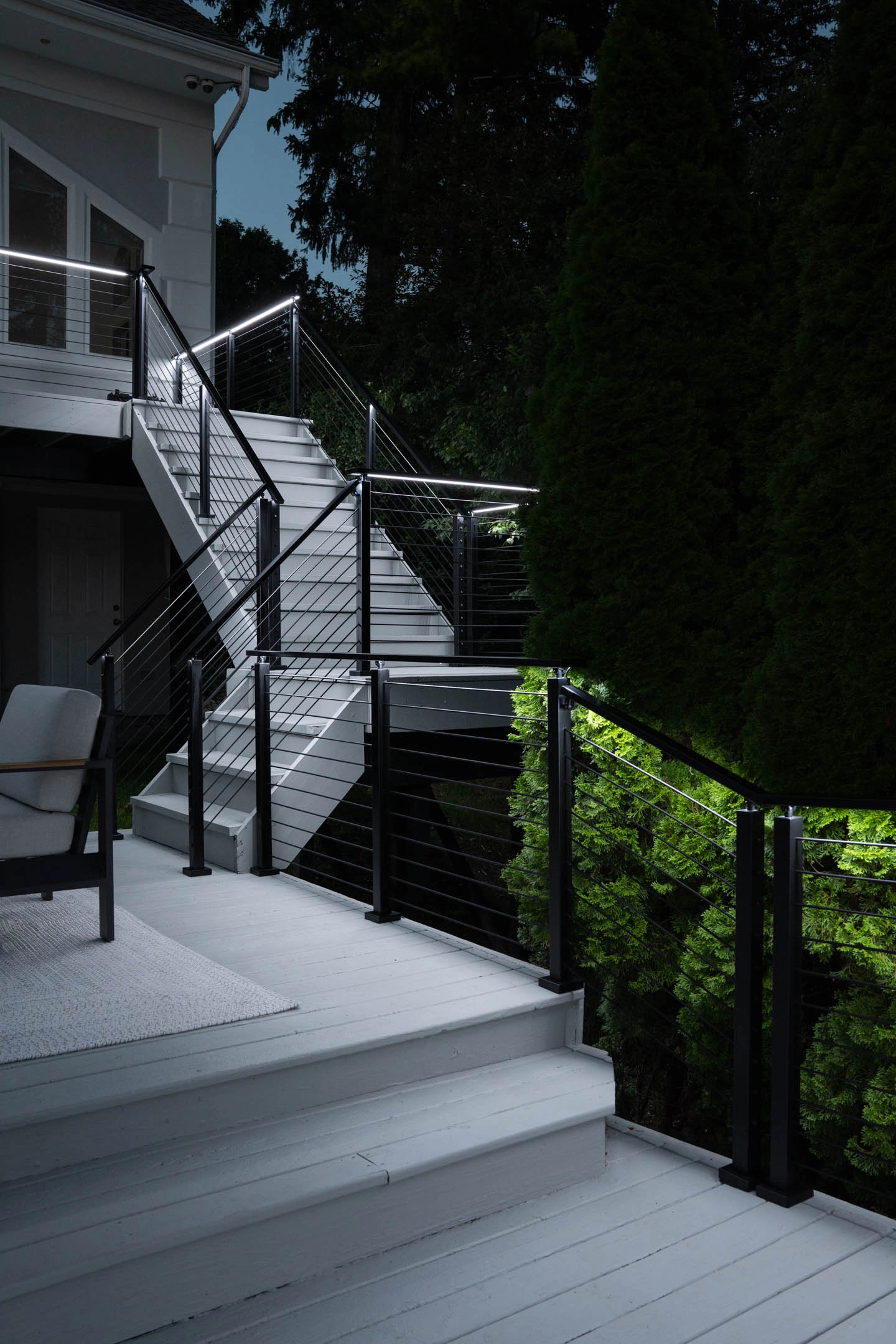 Deck and stair after dark with lit rails beside a tall hedge