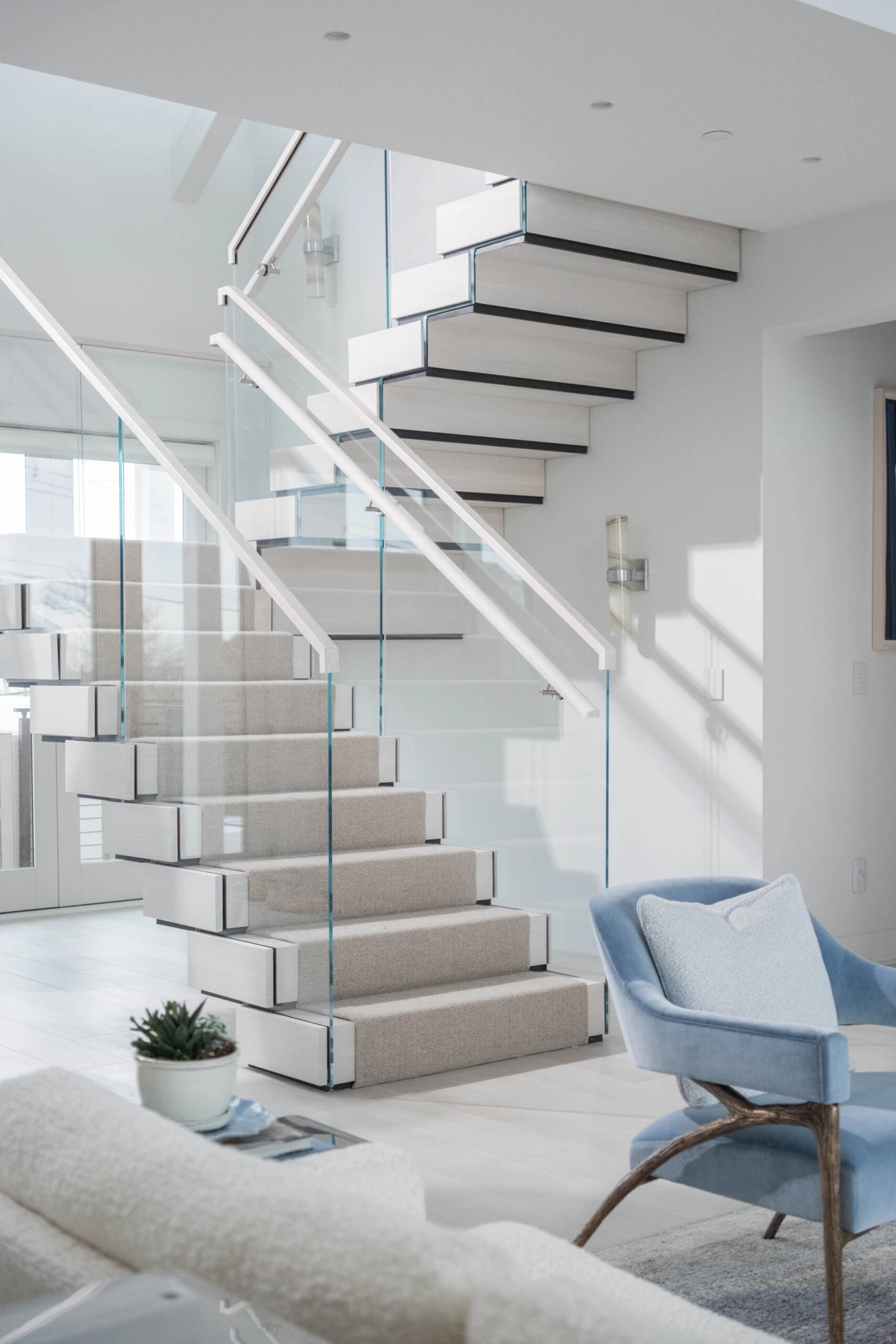 Stair with glass railing beside a pale blue armchair in an open living space