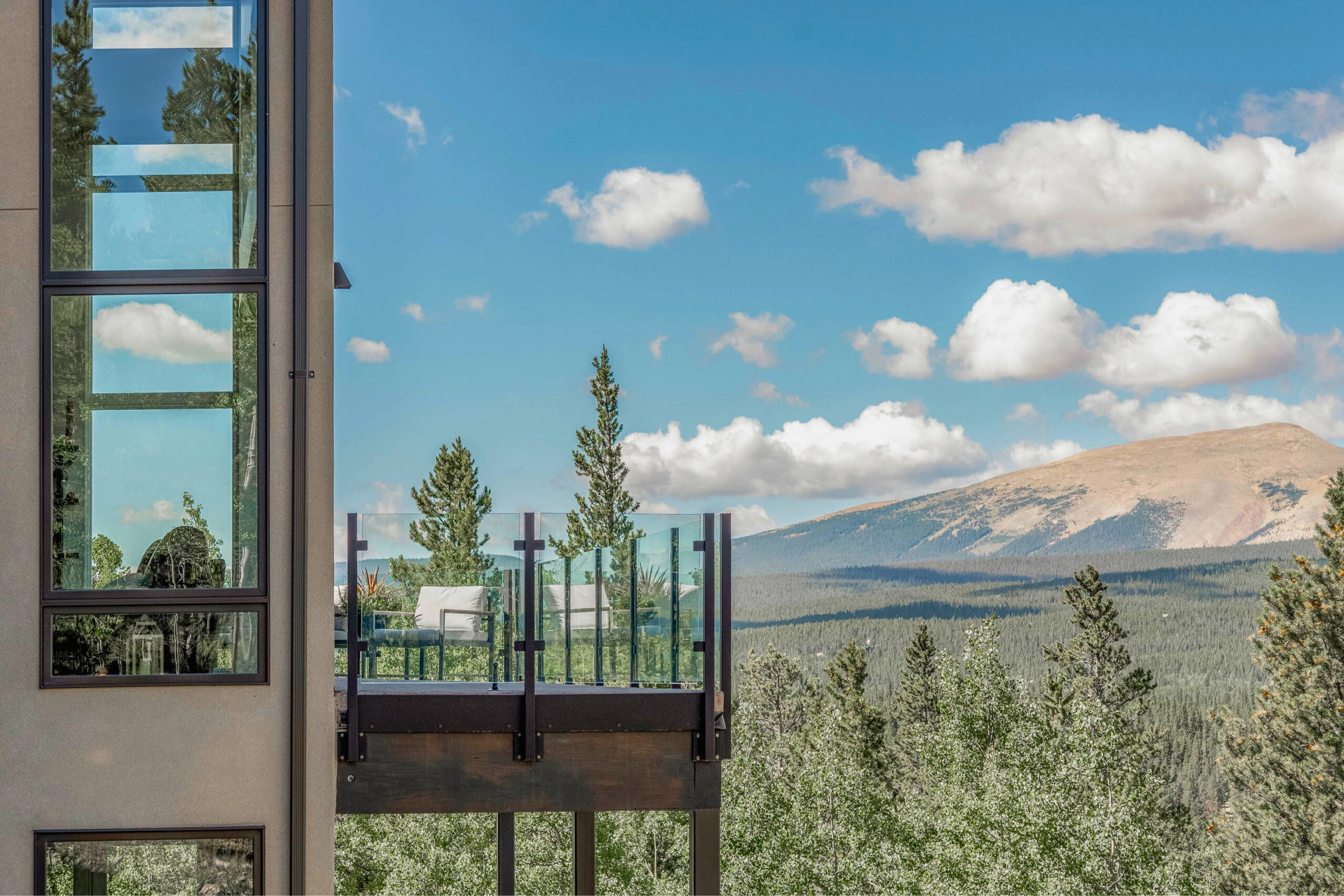 Glass-paneled deck railing on a hillside house framed by pines and a distant peak