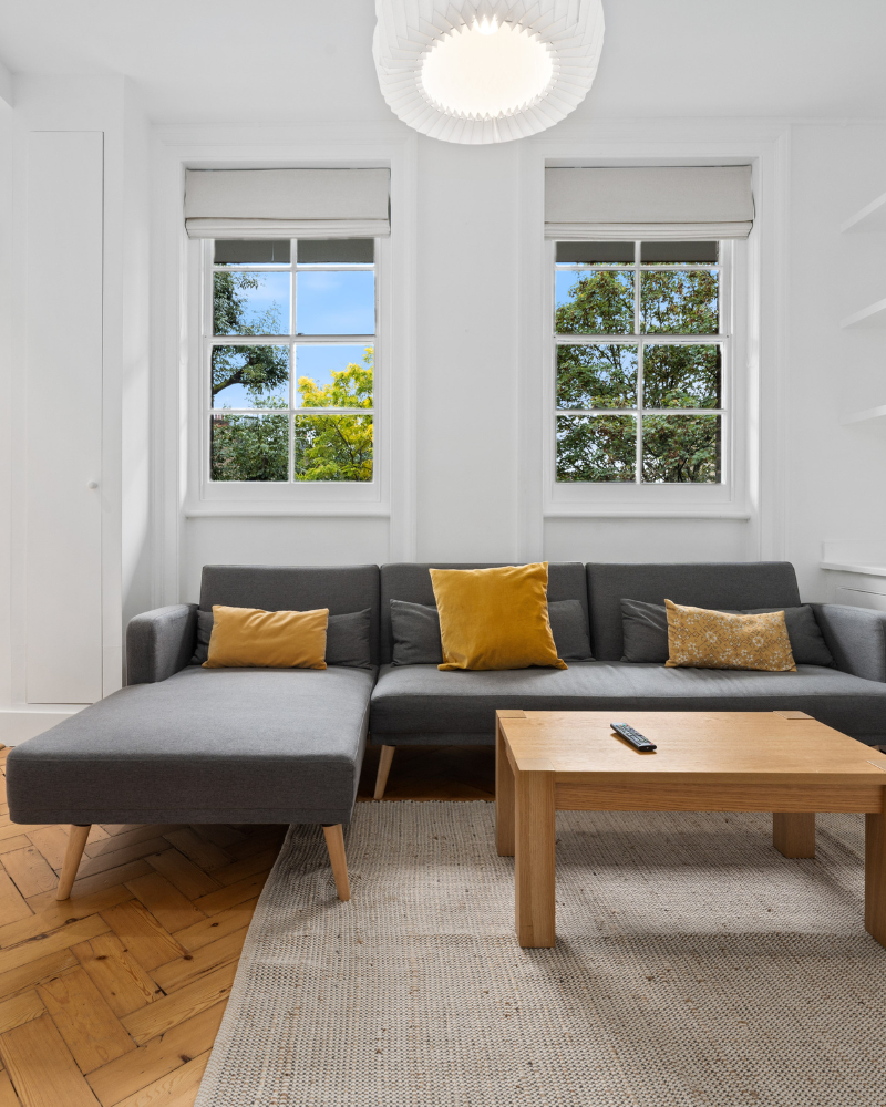 A cozy interior shot shows a black leather sofa next to a light wood open-shelf bench with a plant and books, below a light wood slatted staircase and a framed bird artwork.