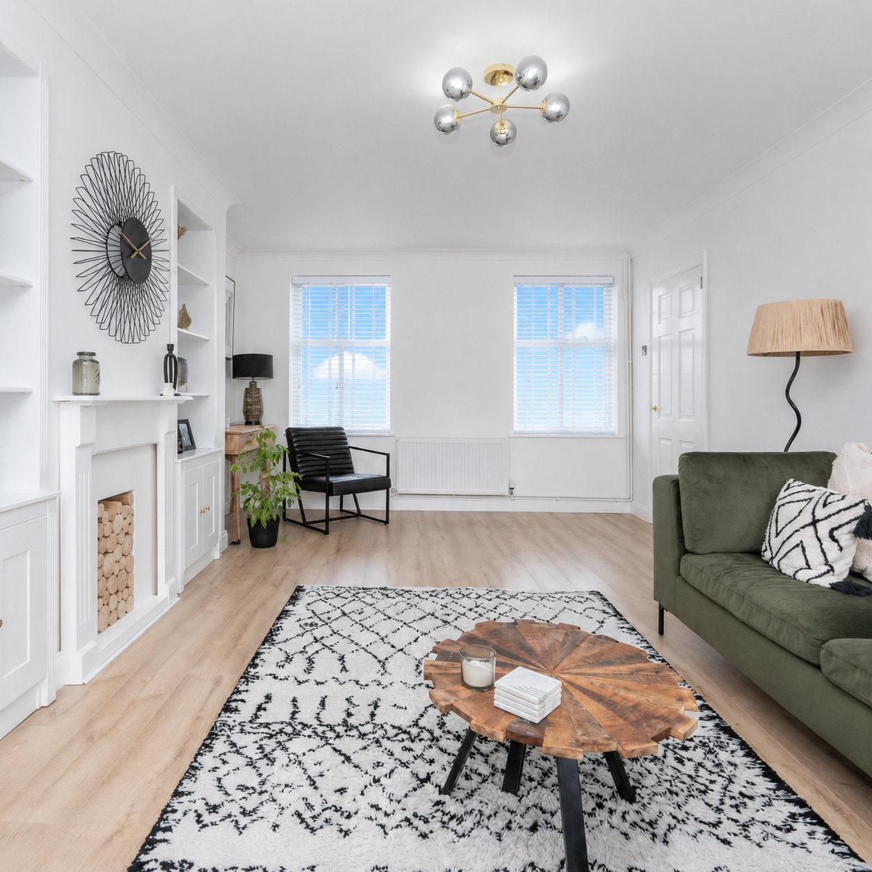 A modern dining room boasts an oval white table with wishbone chairs, a large round mirror above a woven buffet, and a vintage-style pendant light, all on a patterned rug.