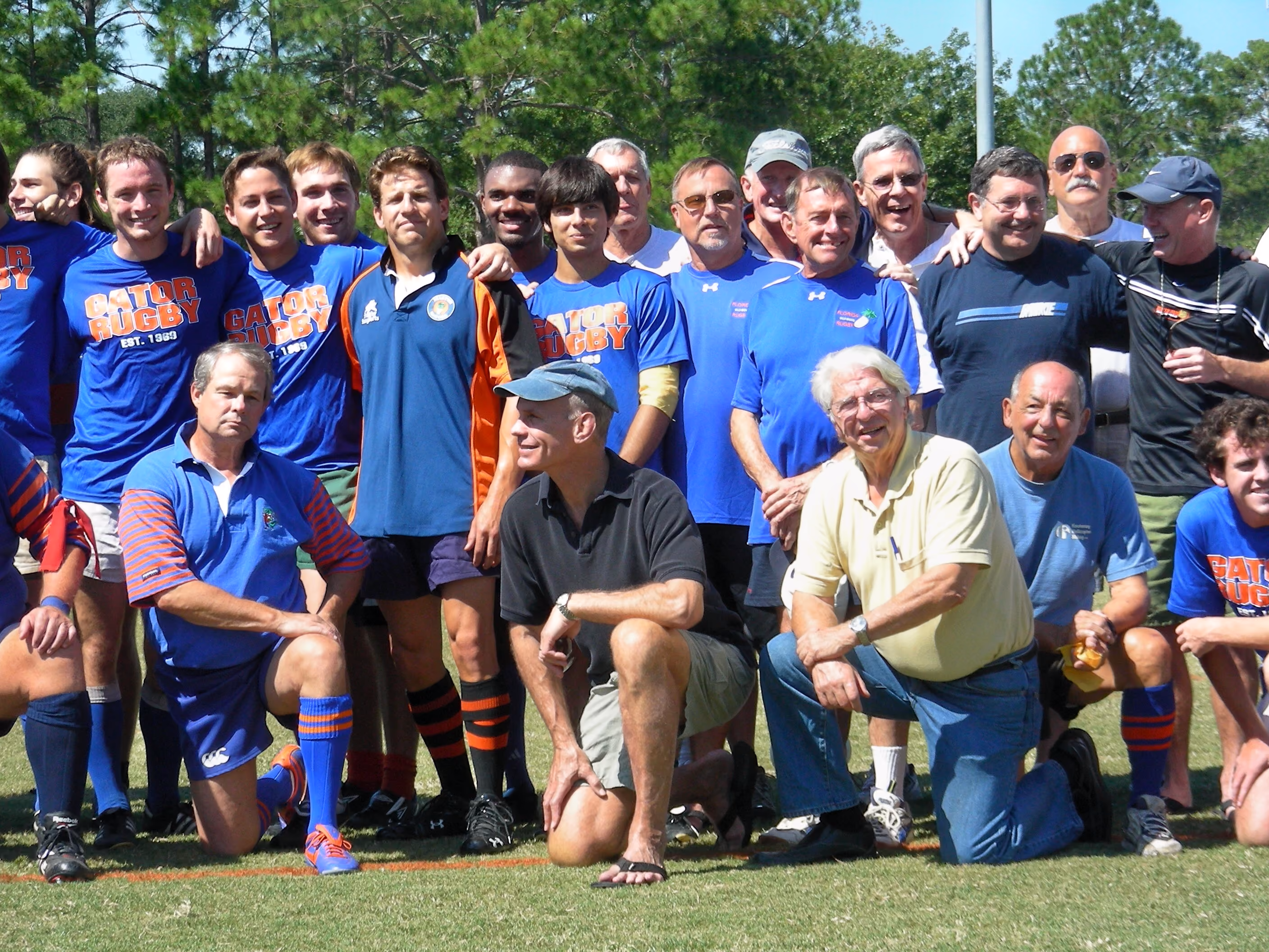Group photo of men on grass field, some in blue 'Gator Rugby' shirts, others in casual clothing.