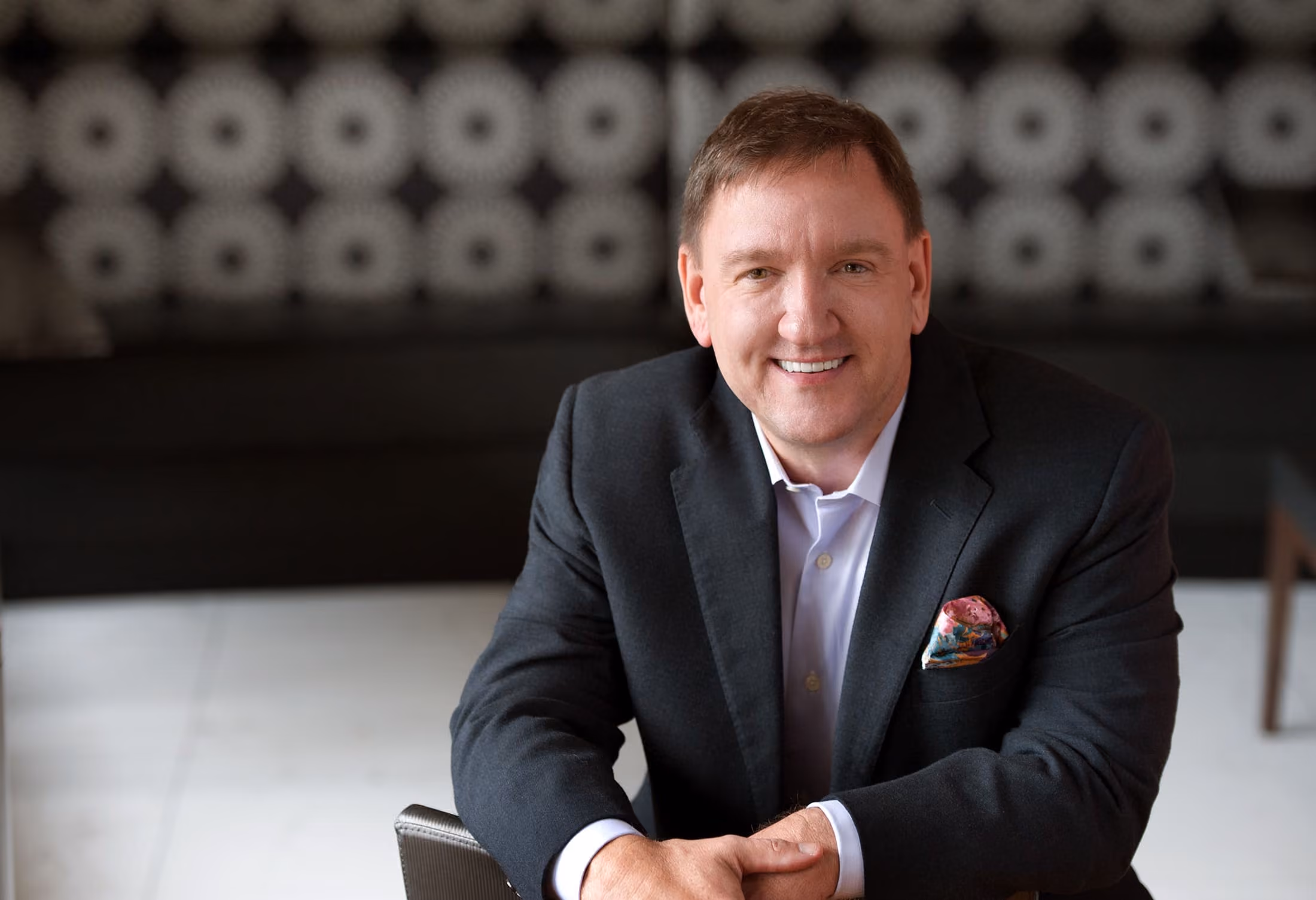 Smiling man in a dark suit jacket with a colorful pocket square, sitting with hands clasped in front of him against a blurred patterned background.