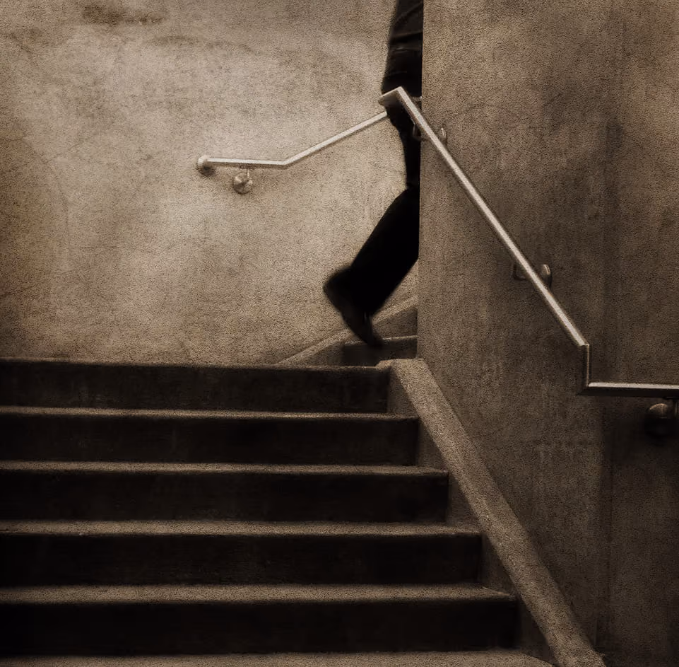 Shadowy image of a person partially visible walking up a dimly lit concrete staircase with metal handrails on both sides.