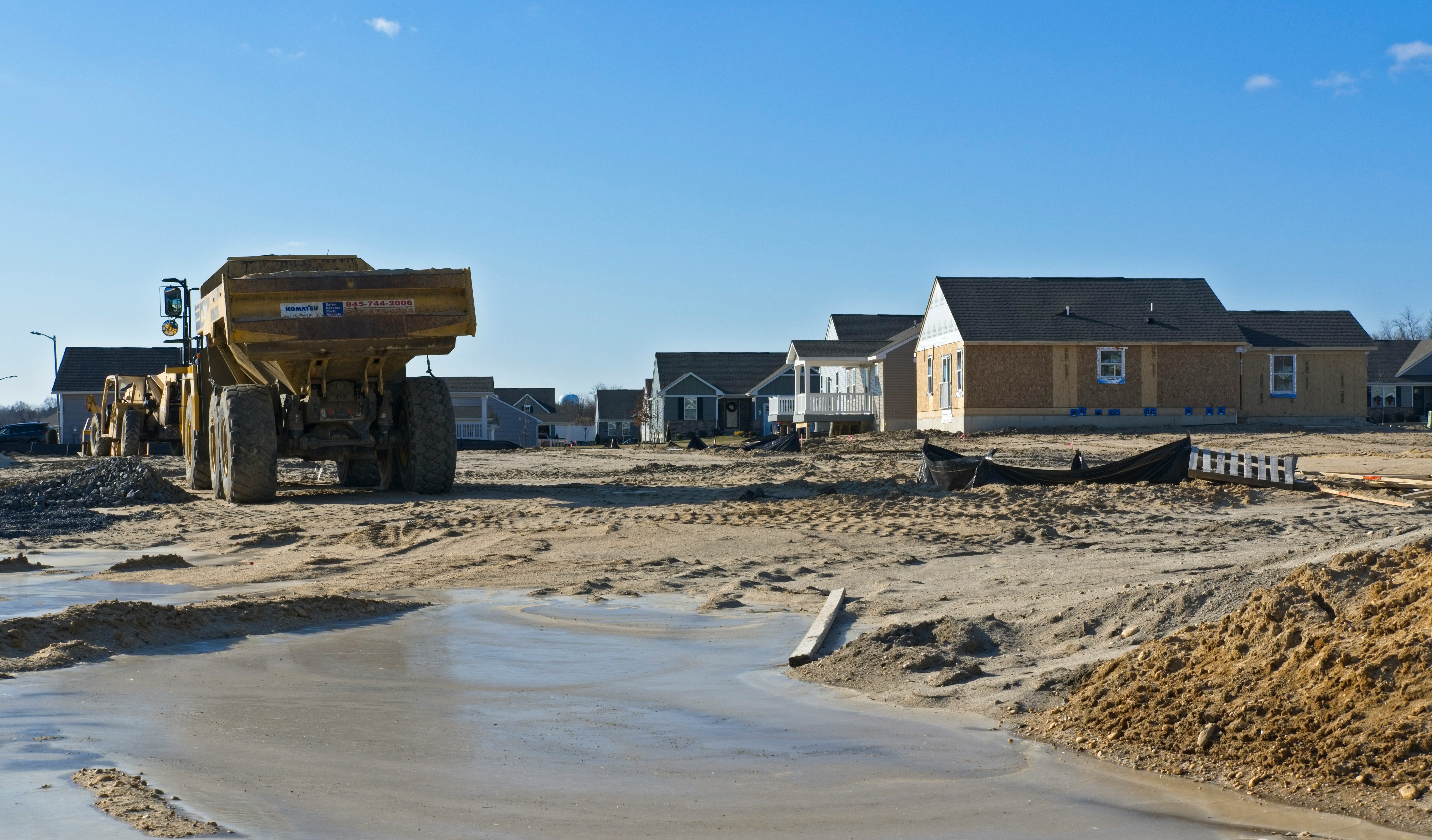 Construction site with a large dump truck parked on sandy ground near partially built houses under a clear blue sky.