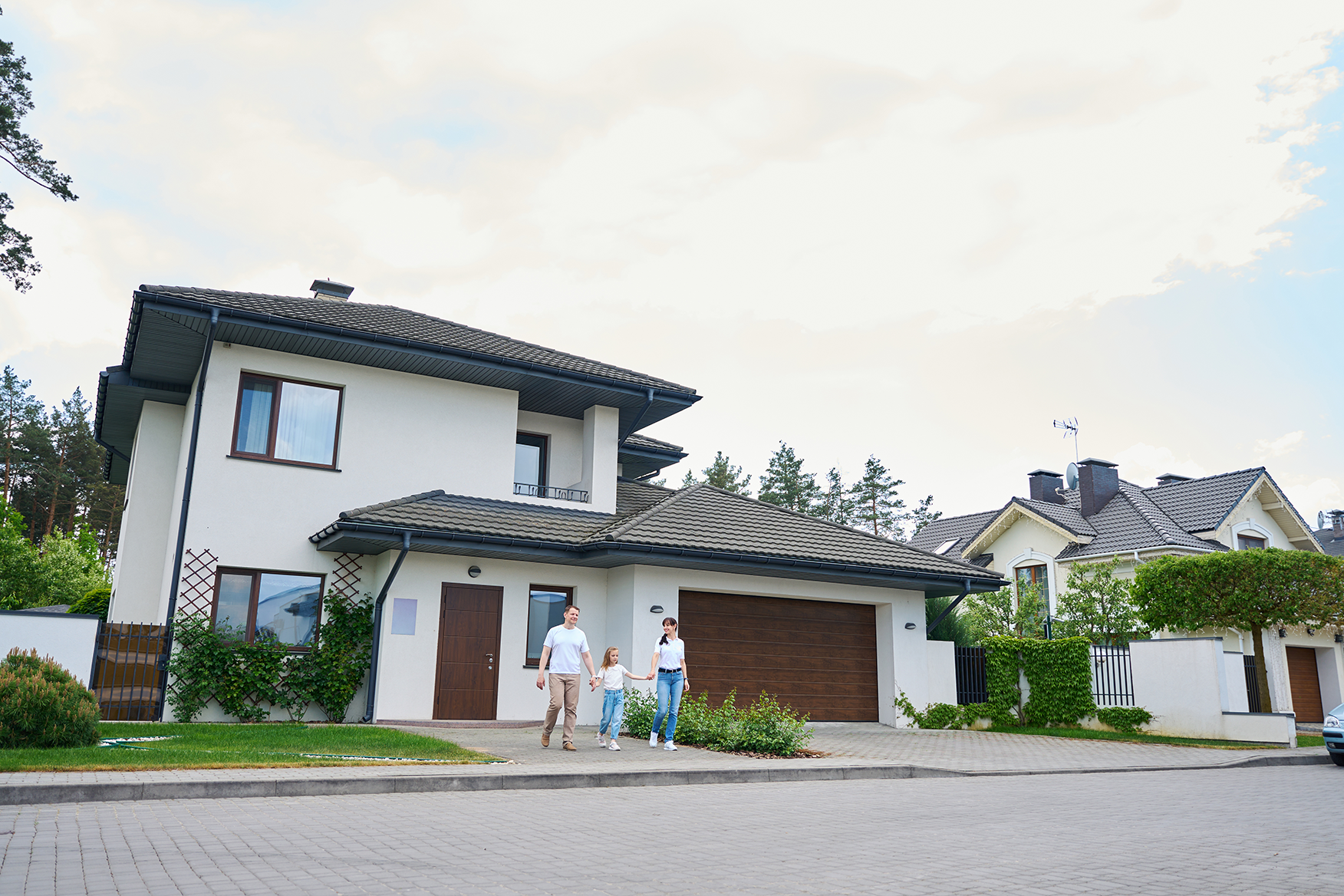 Family of three holding hands and walking on the driveway of a modern two-story house with a double garage.