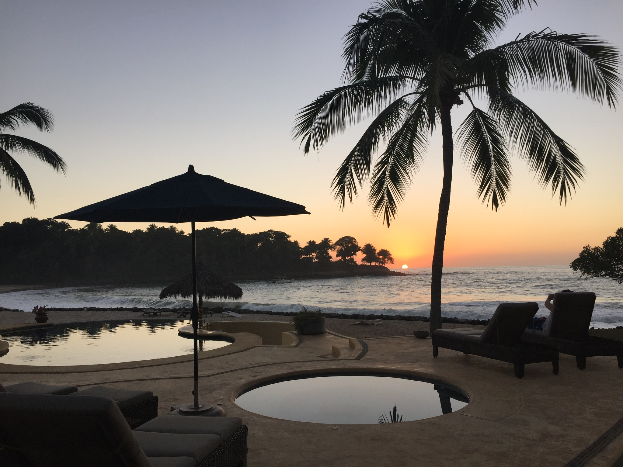 Sunset over the ocean framed by palm trees, with a beachfront pool, lounge chairs, and umbrella in the foreground.