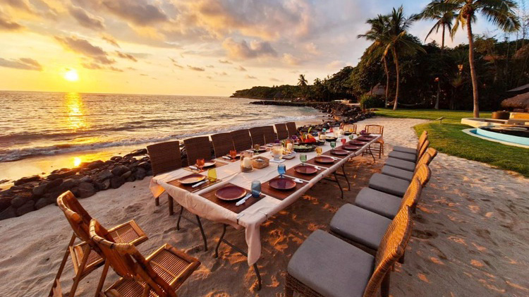 A beachfront dining setup features a long table with various dishes and colorful drinks. Surrounding the table are several chairs, with tropical palm trees and the ocean visible in the background during sunset.