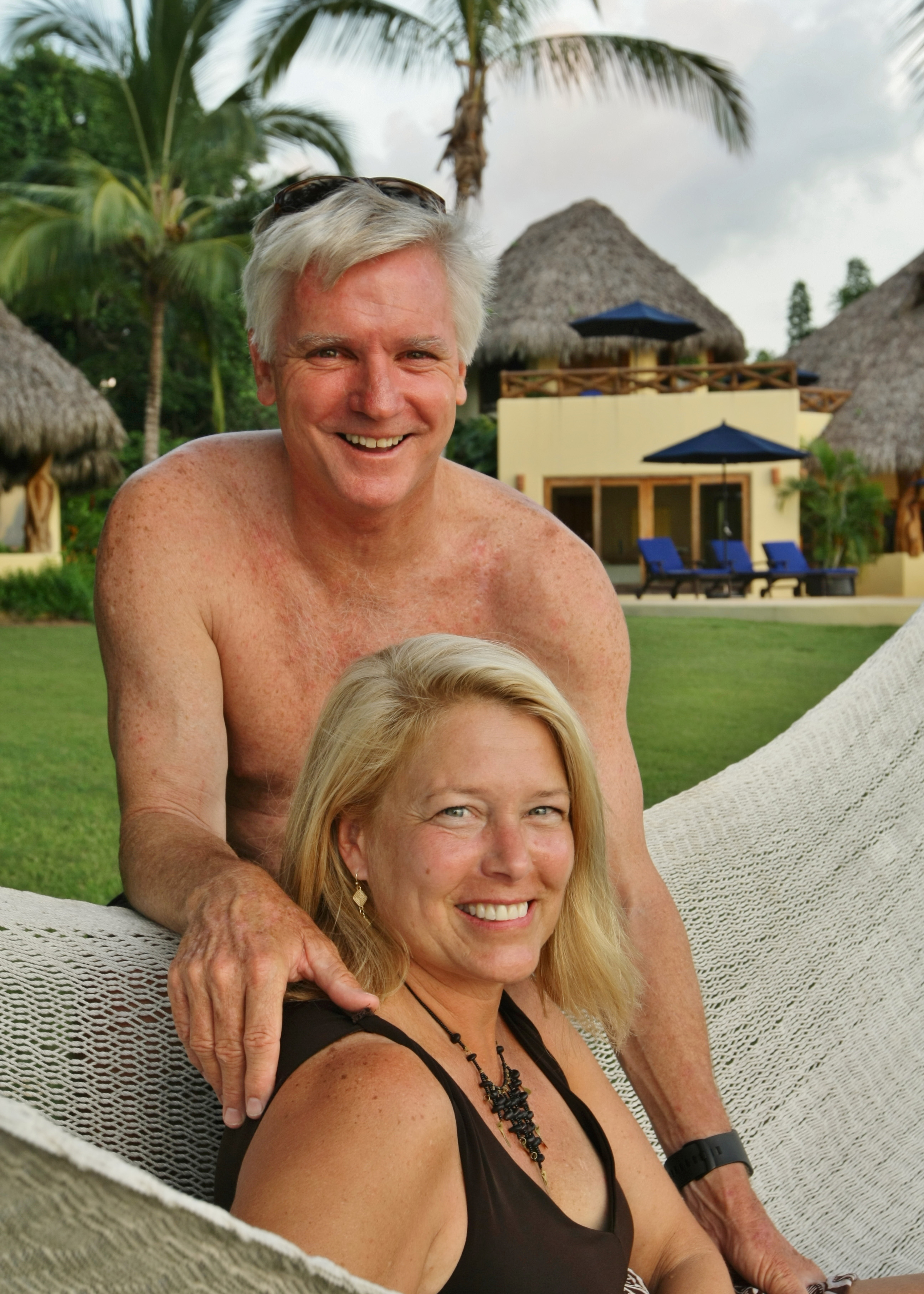 Smiling couple relaxes together in a hammock at a tropical resort with palm trees and thatched-roof villas in the background.