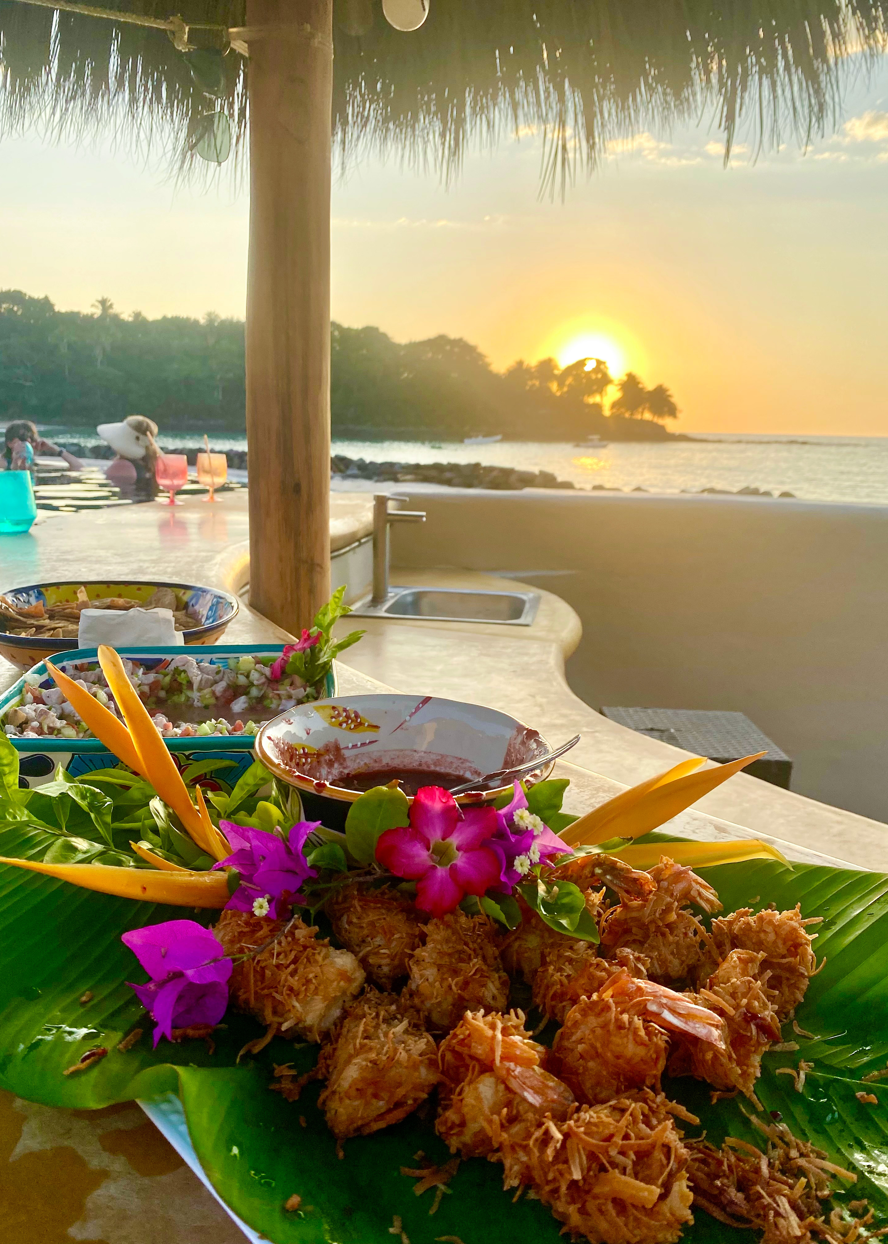 Coconut-crusted shrimp served on a banana leaf at an open-air restaurant overlooking the ocean during sunset.