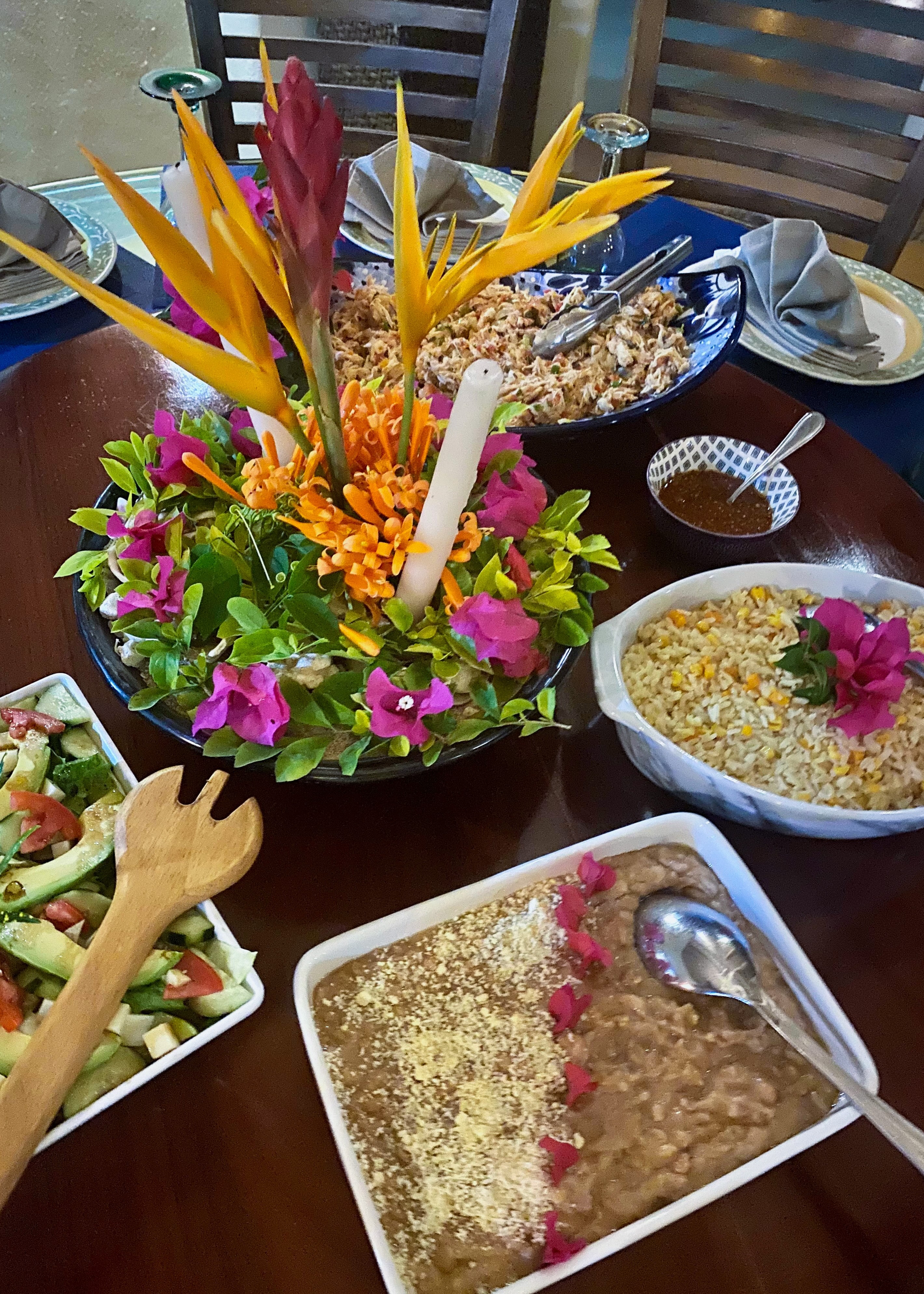 Table set with a tropical centerpiece of bird of paradise flowers and bougainvillea, surrounded by dishes of refried beans, corn rice, crab salad, cucumber-tomato salad, and a side of dipping sauce.