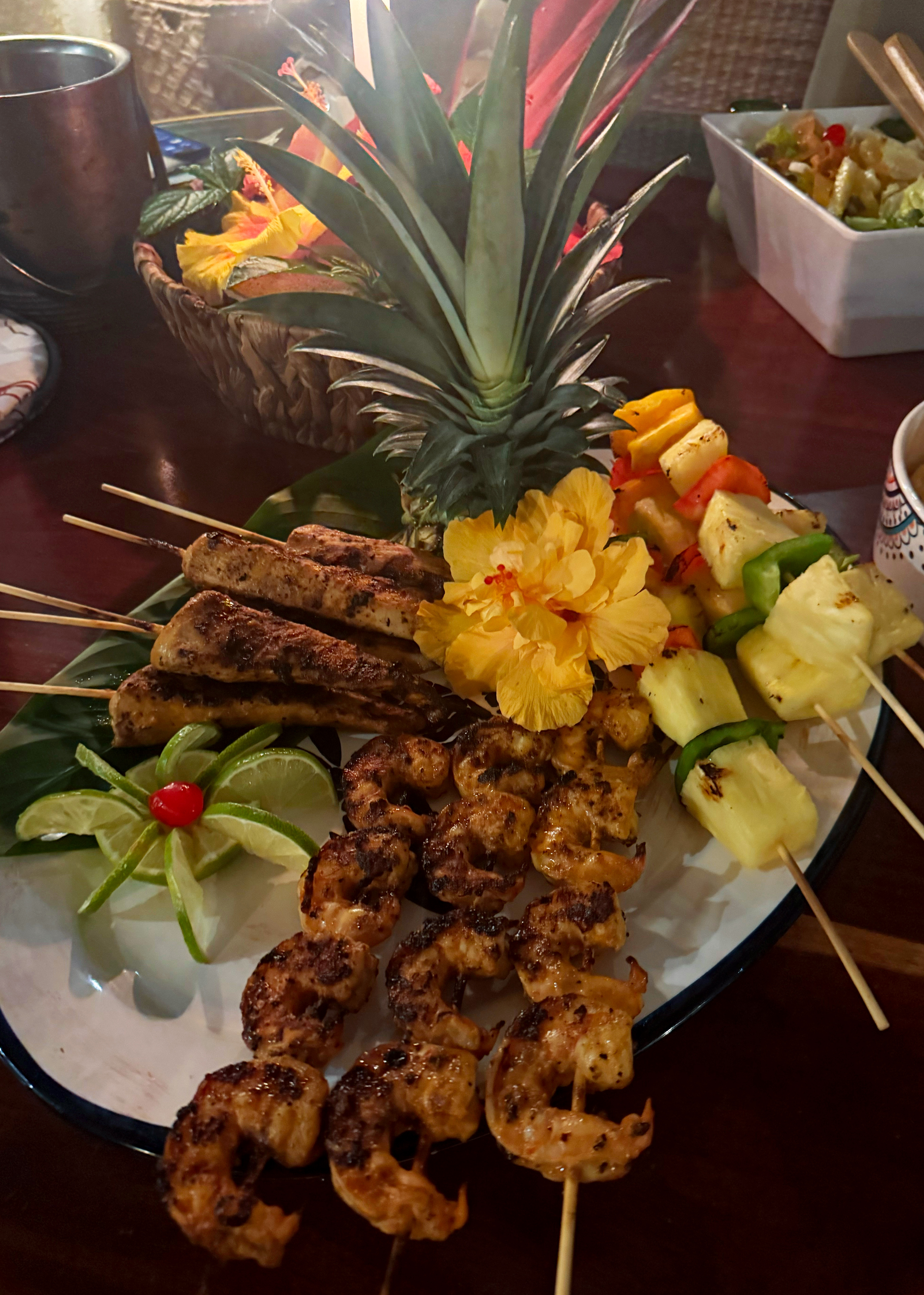 "Plate of grilled shrimp and chicken skewers with pineapple and bell pepper kebabs, garnished with a hibiscus flower, lime wheels, and a whole pineapple, set on a table with a fruit basket and side salad in the background.
