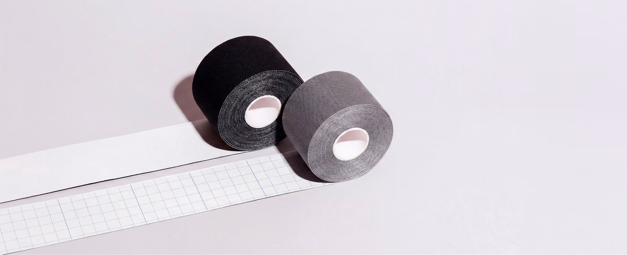 Zwei Rollen Klebeband in Schwarz und Grau mit entrolltem, kariertem Trägerpapier auf hellgrauem Hintergrund.