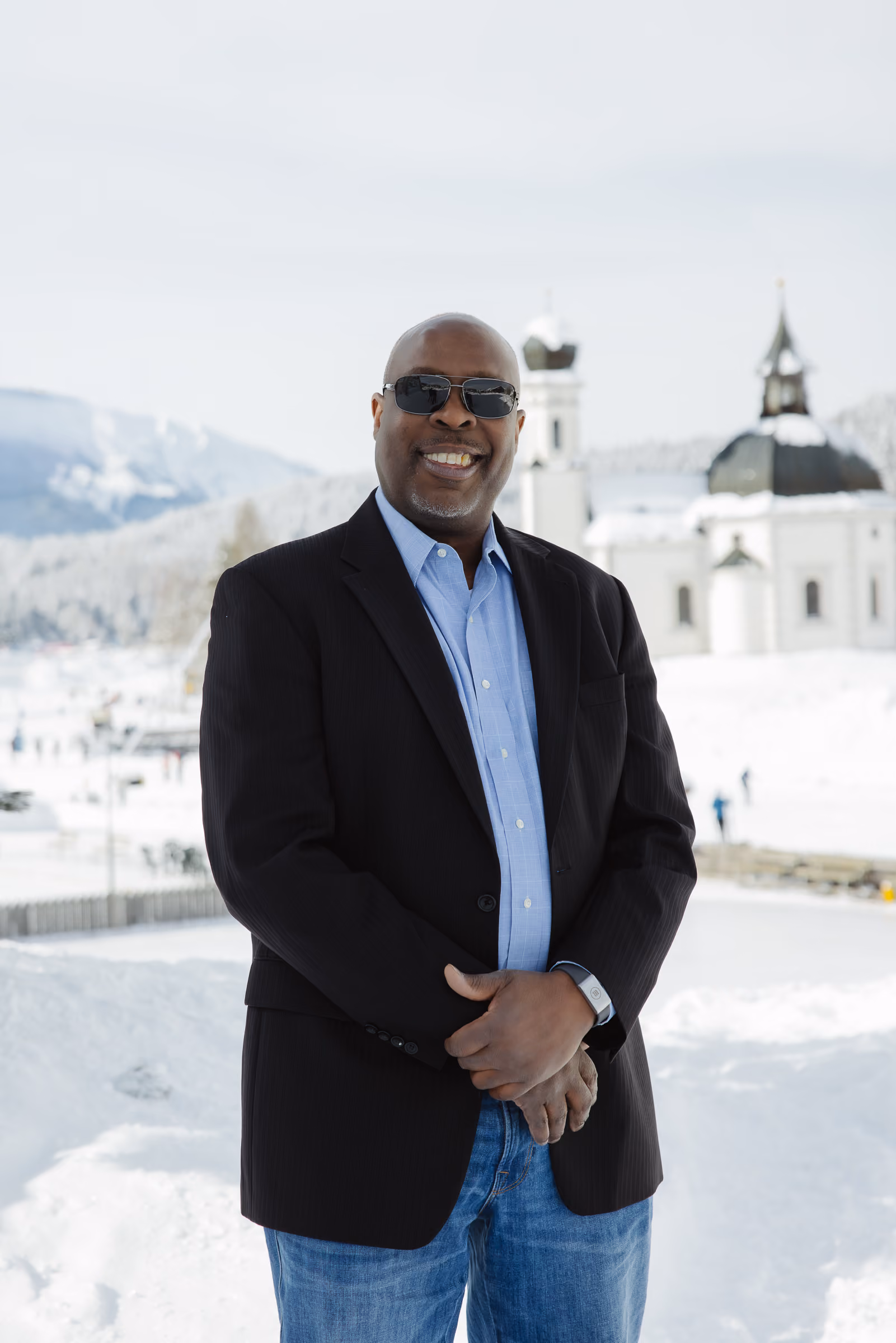 Mann in schwarzem Sakko und blauer Hemdjacke steht vor verschneiter Kirche und Bergen.