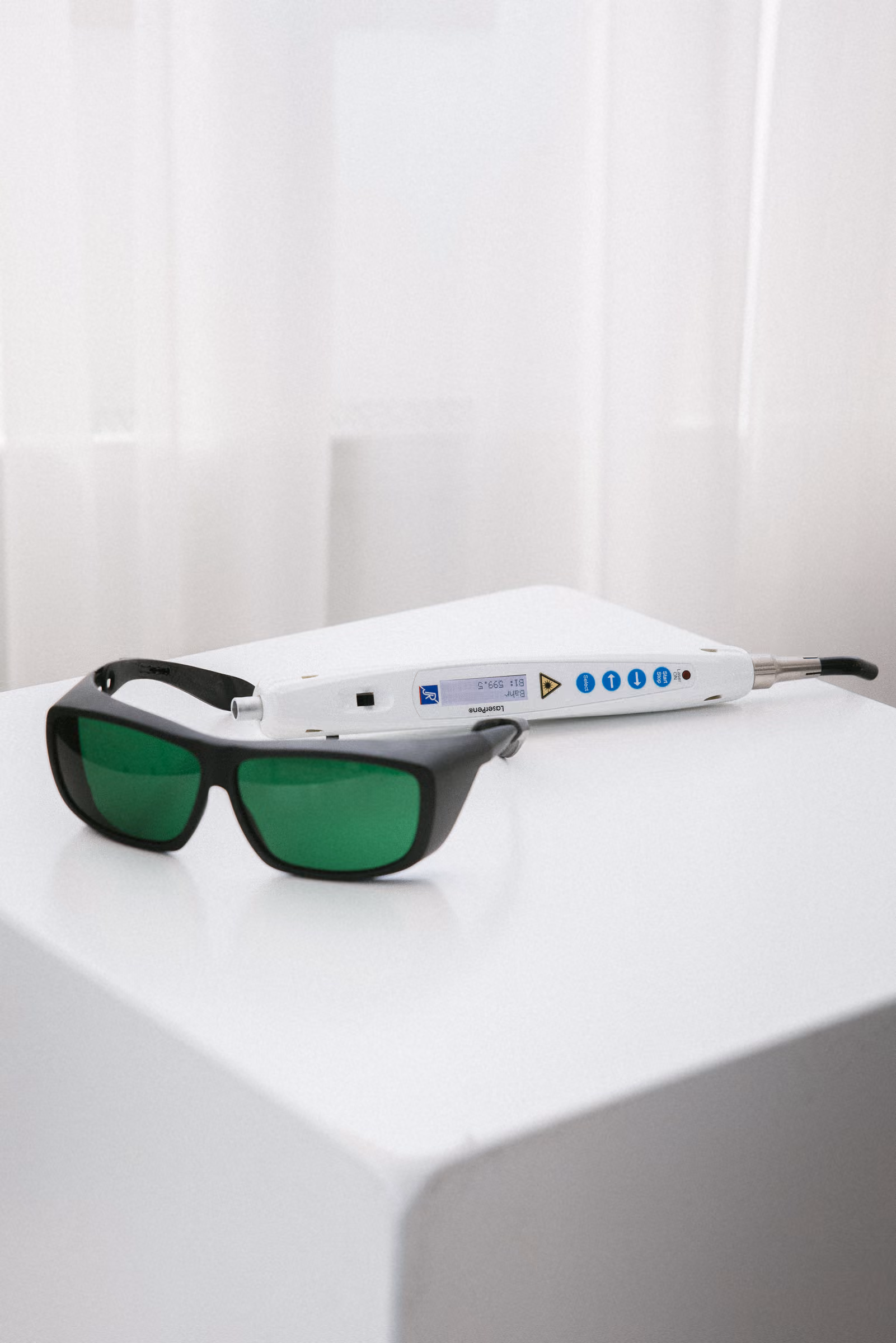 Schwarze Schutzbrille mit grünen Gläsern neben einem weißen Lasergerät auf weißem Tisch.