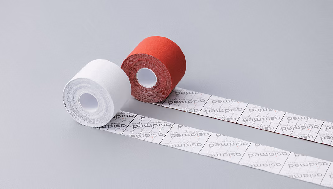 Zwei Rollen Kinesiologie-Tape, eine weiß und eine rot, mit jeweils ausgeschnittenen, länglichen Klebestreifen auf hellem Hintergrund.