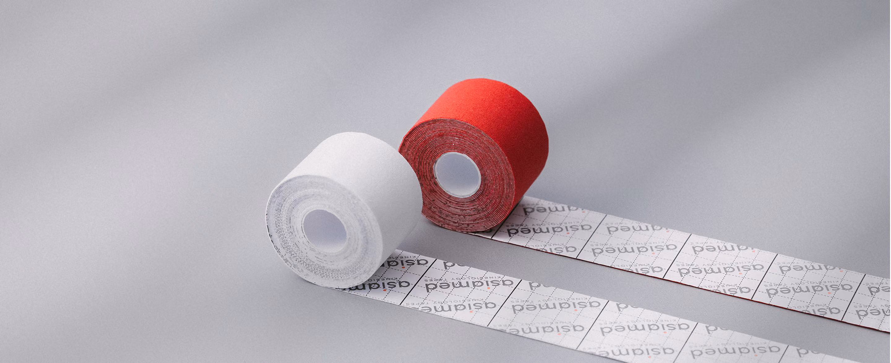 Zwei Rollen Kinesiologie-Tape, eine rot und eine weiß, mit jeweils einem Teil des Tapes ausgelegt, auf hellem Untergrund.