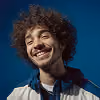 Smiling young man with curly hair and beard wearing a light jacket over a white shirt.