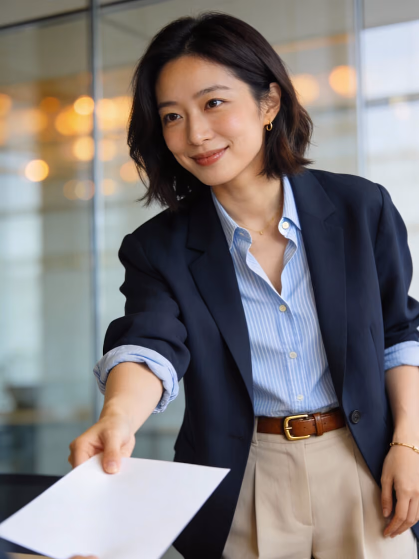 Smiling professional woman in blazer handing over a white sheet of paper in an office setting.