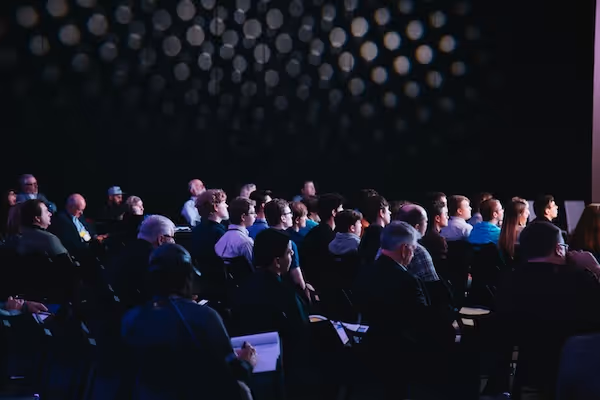 Audience sitting in rows watching a presentation in a darkened room with spotlight patterns on the wall.