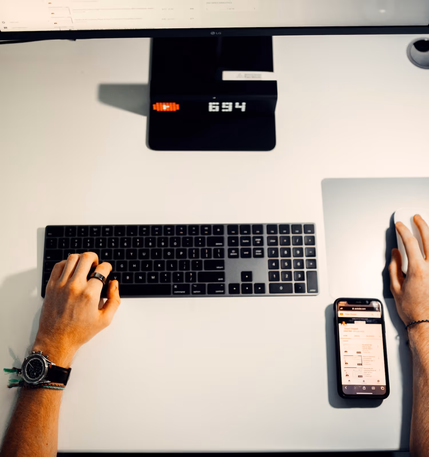 Person typing on keyboard and using a mouse next to a smartphone on a white desk with a monitor above.