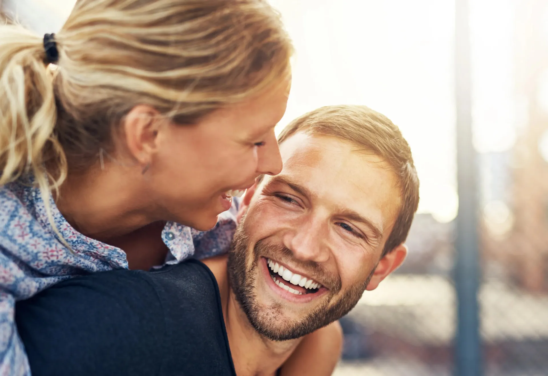 Smiling woman leaning on a man's back, both sharing a joyful moment outdoors.