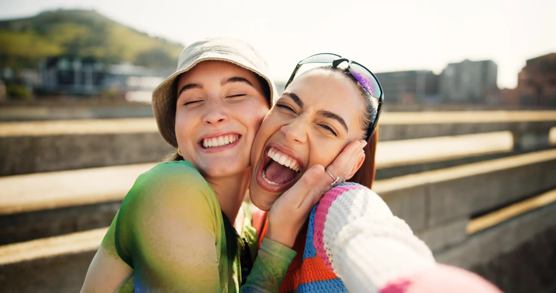 Two young women smiling and hugging closely outdoors while taking a selfie on a sunny day.