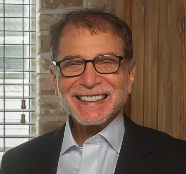 Smiling older man with glasses, gray beard, and dark suit jacket in a warmly lit room.