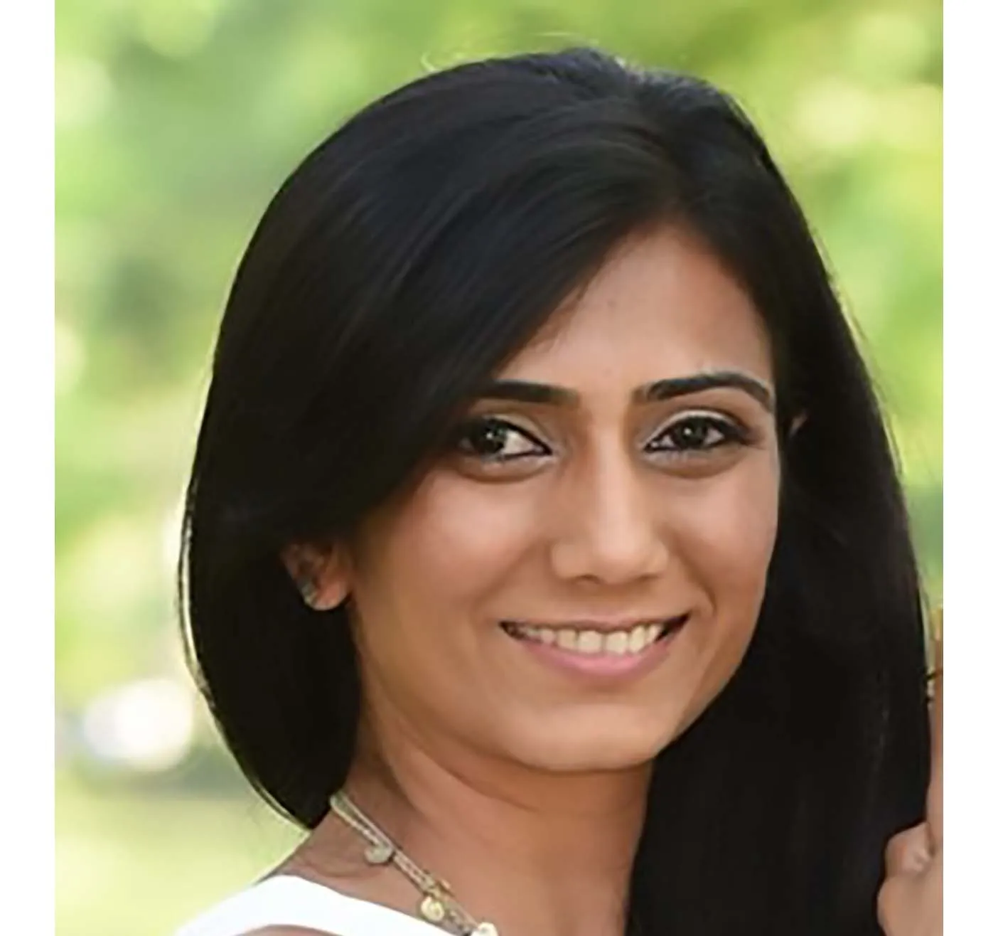 Portrait of a smiling woman with long black hair and a necklace in an outdoor setting.
