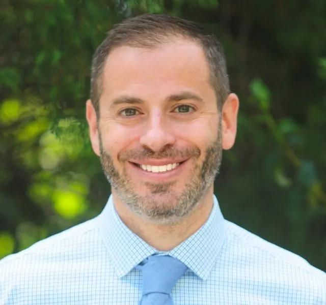 Smiling man with short hair and beard wearing light blue shirt and blue tie outdoors.
