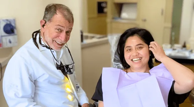 Smiling male dentist in white coat with dental magnifier and female patient in dental chair.