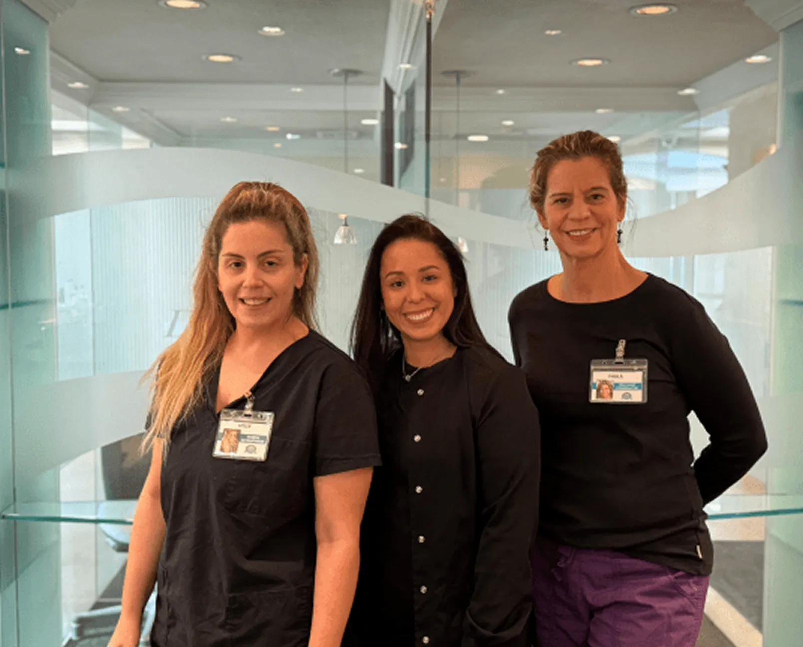 Three women wearing black medical scrubs with ID badges standing inside a modern office.