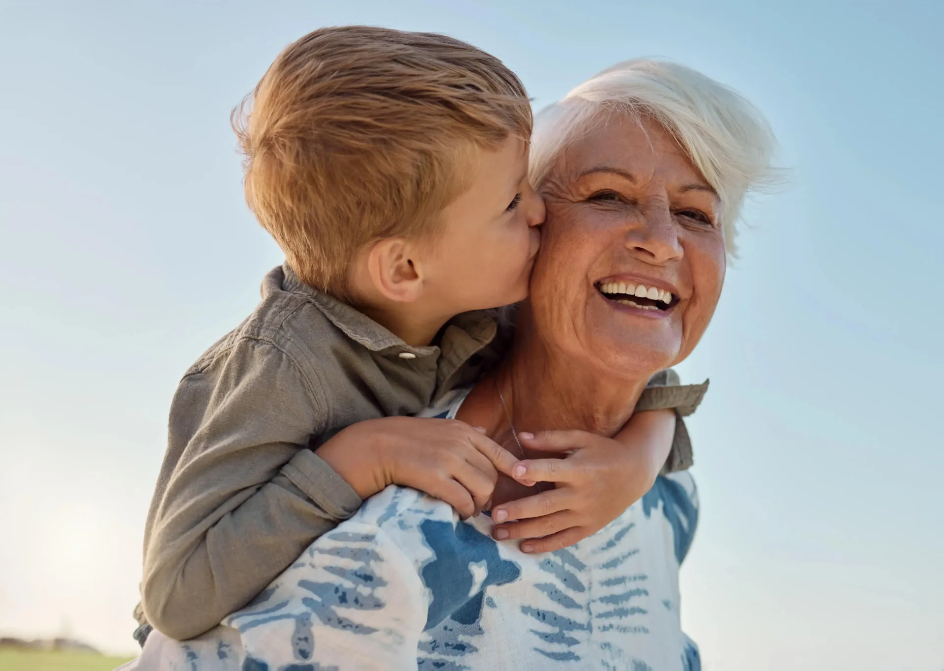 Young boy kisses smiling elderly woman on cheek outdoors under clear sky.
