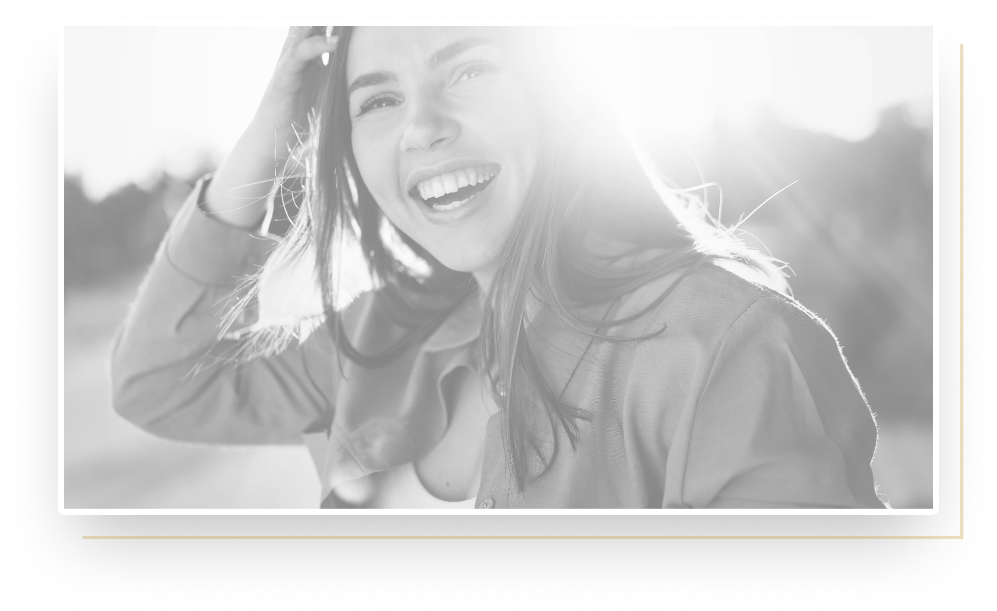 Close-up of a smiling woman with long hair touching her head in bright sunlight outdoors.