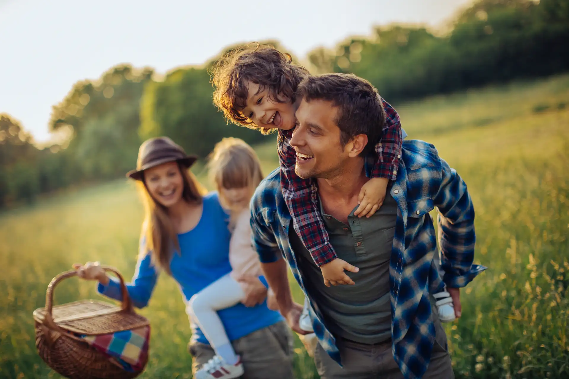 Smiling family enjoying outdoor picnic with children on the father's back in a sunny green field.