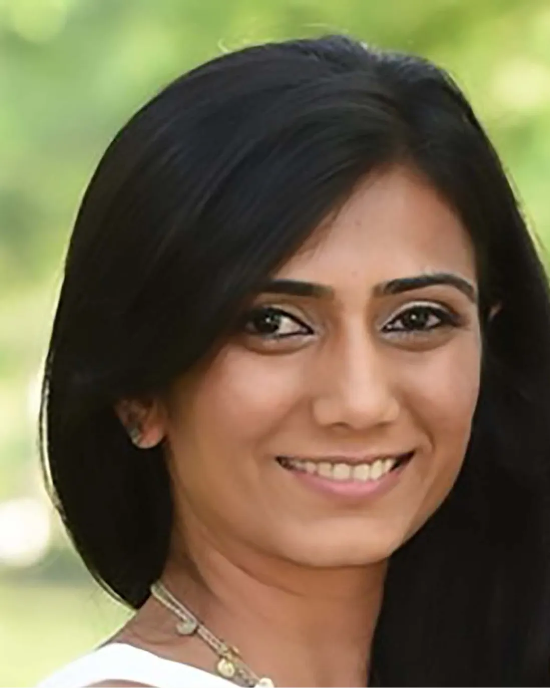 Smiling woman with long black hair and a necklace against a blurred green background.