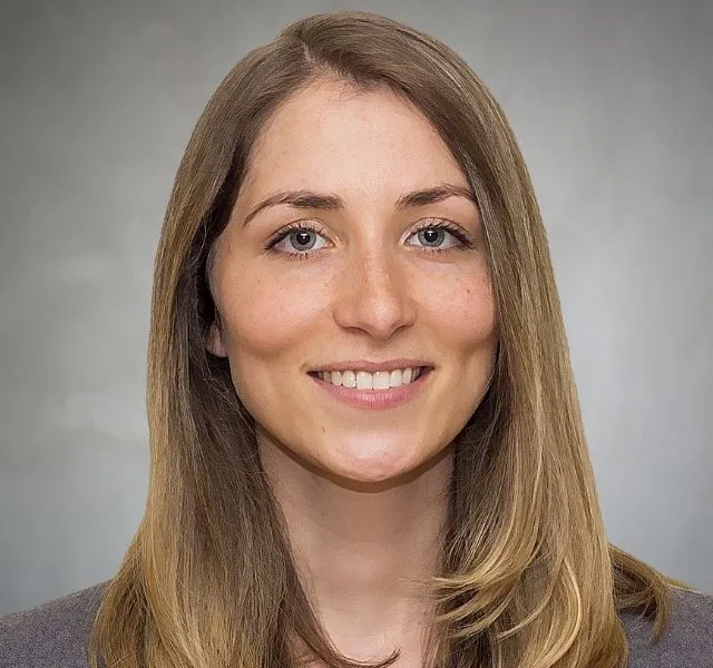 Headshot of a young woman with long light brown hair smiling against a gray background.