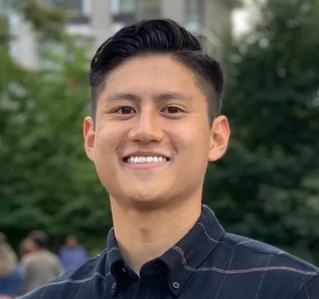 Smiling young man with dark hair wearing a dark button-up shirt outdoors with blurred background.