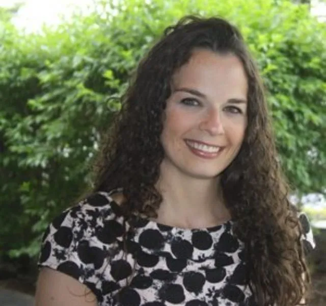Smiling woman with long curly hair wearing a black and white patterned top outdoors with greenery behind her.
