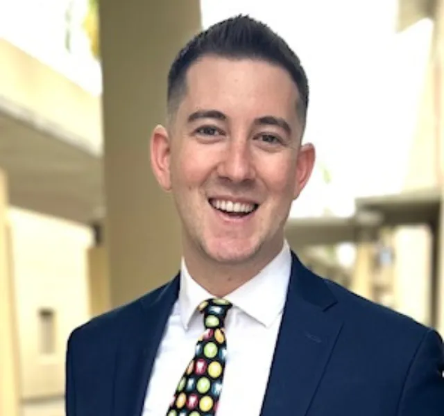 Smiling man in navy suit and colorful polka dot tie standing outdoors in a bright, blurred background.