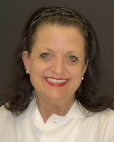 Smiling woman with dark hair, red lipstick, and pearl earrings wearing a white collared shirt.