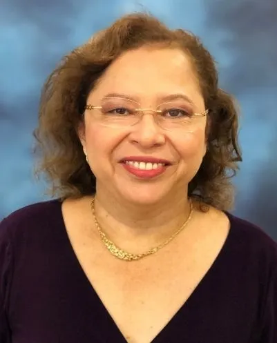 Smiling woman with glasses and curly hair wearing a gold necklace and dark purple top on a blue background.